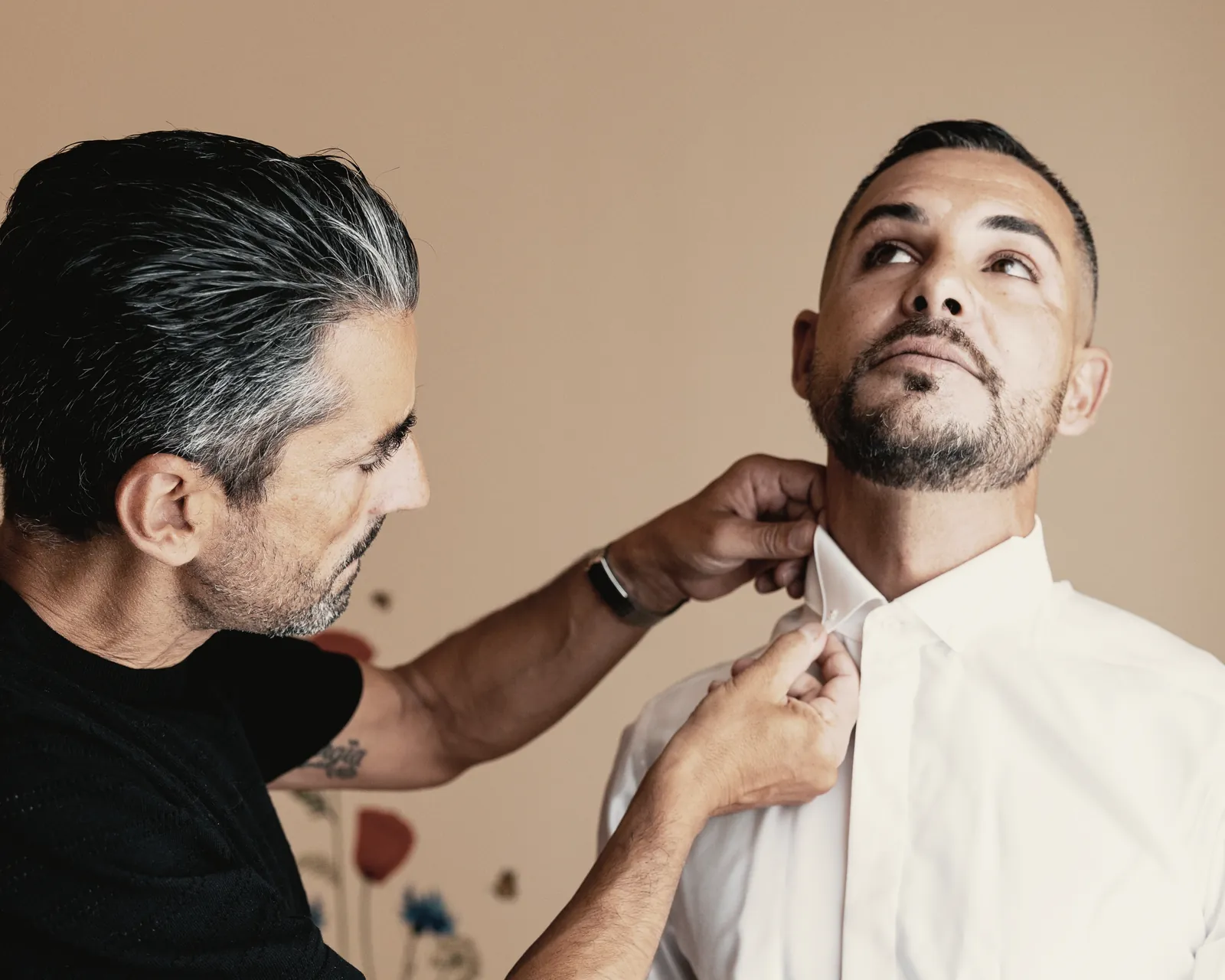 Two hands fasten the groom’s shirt collar as he lifts his chin to the warm light of the room.