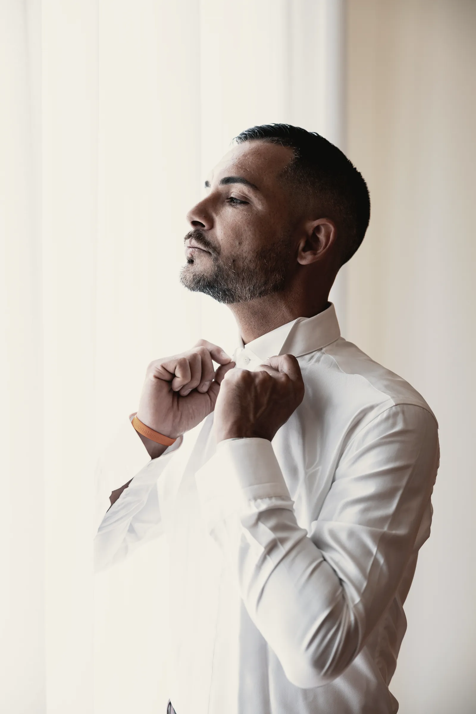 The groom by the pale curtain, eyes closed, fastening his collar.