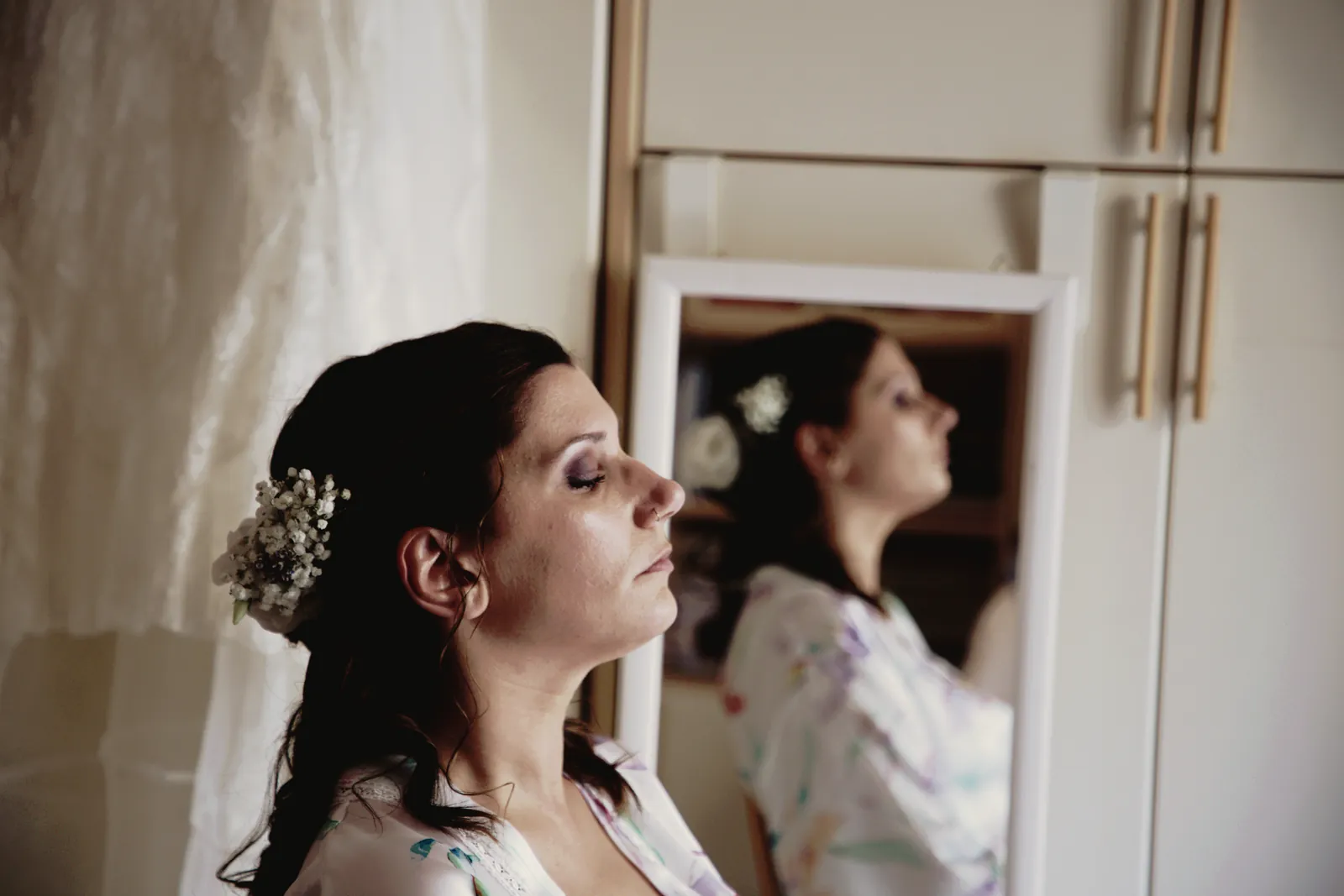 La sposa a occhi chiusi, il viso alzato verso la luce; nello specchio, il suo profilo.