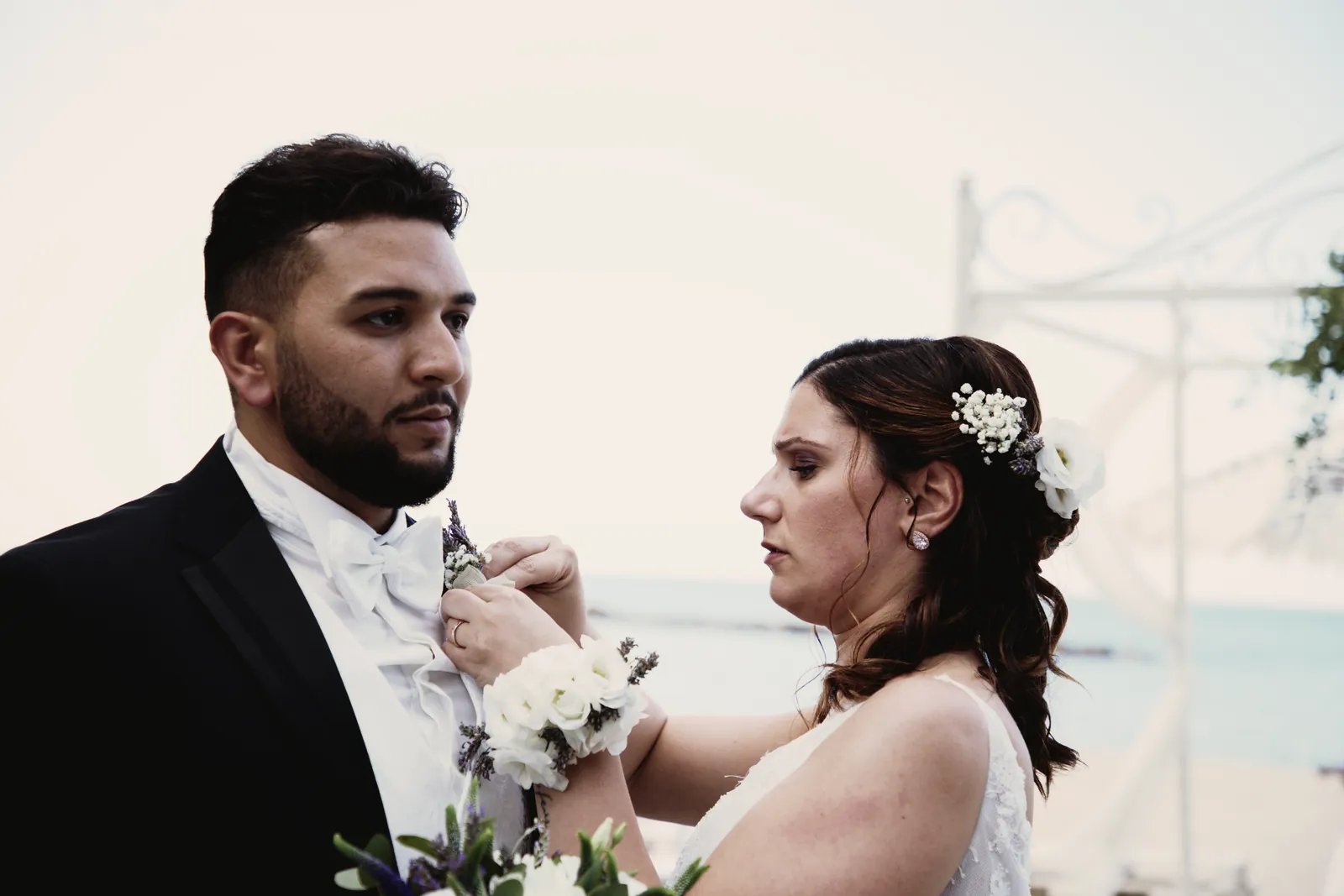La sposa sistema il fiore all’occhiello dello sposo; dietro di loro, il mare.