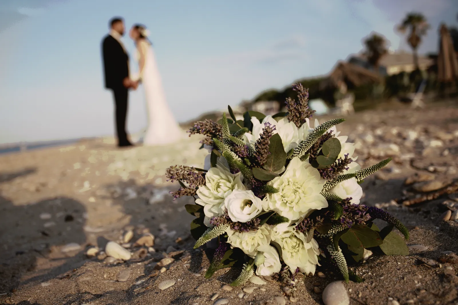 Il bouquet posato sulla sabbia fra le conchiglie; dietro, sfocati, gli sposi mano nella mano.