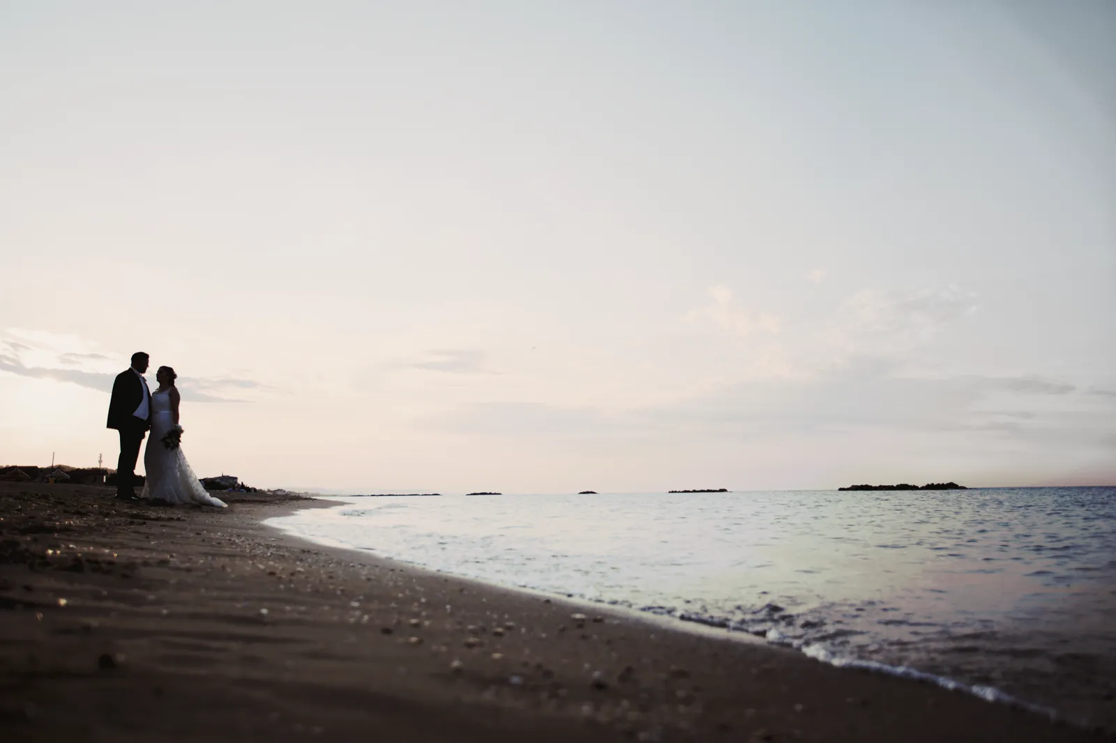 The couple in silhouette at the water’s edge at dusk, a calm sea and a wide pale sky.