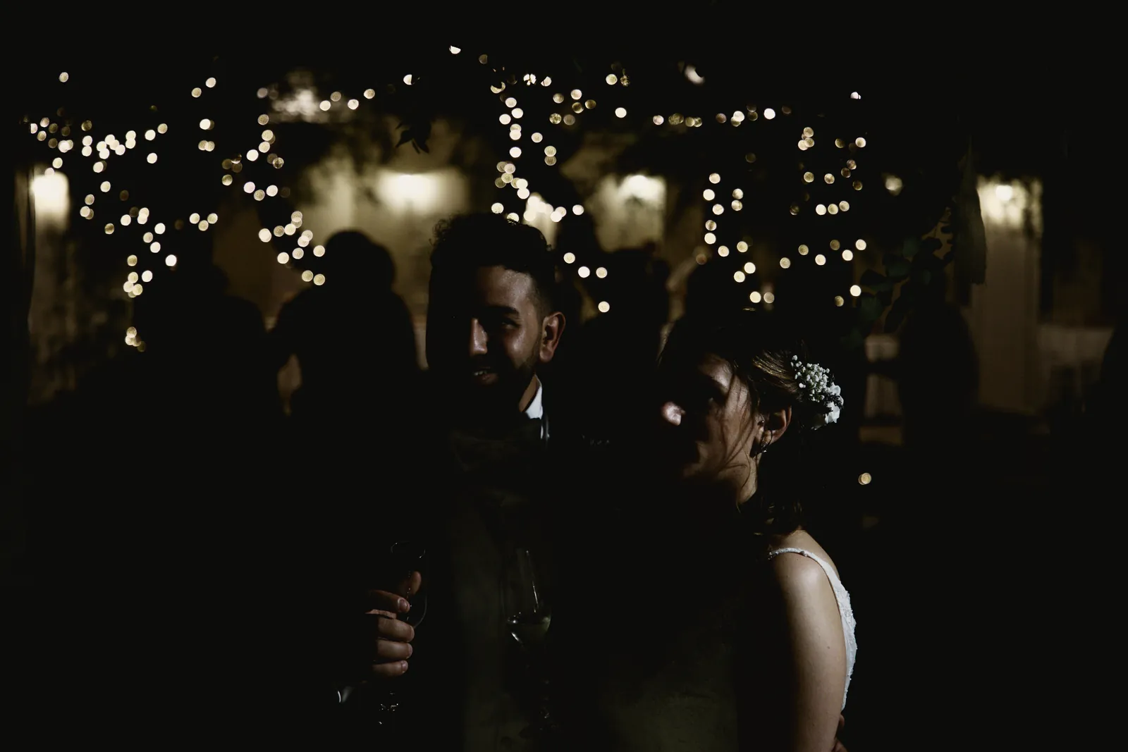 Nel buio, fra le luci del giardino, lo sposo sorride con il calice in mano accanto alla sposa.