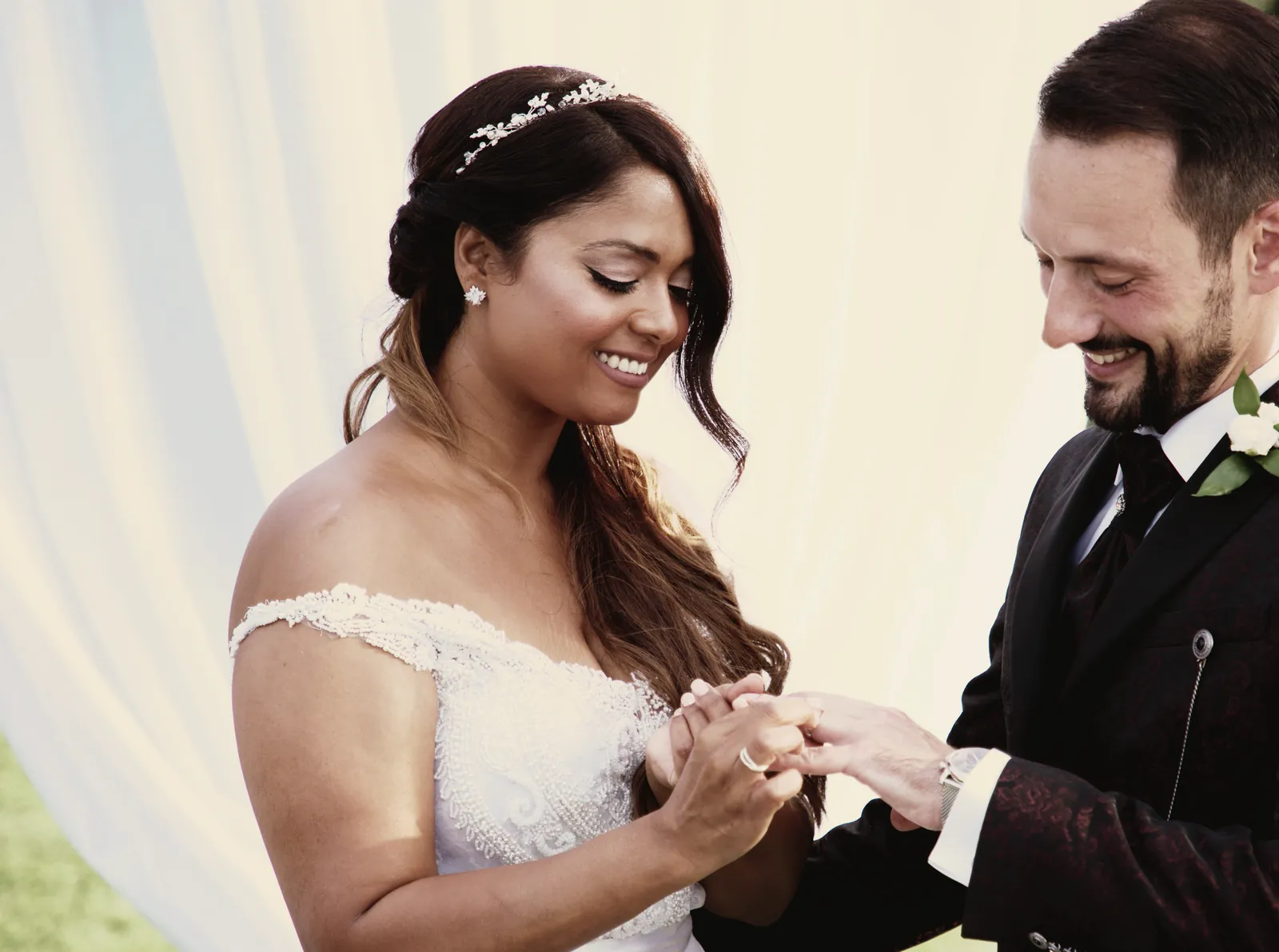 Lo scambio degli anelli: la sposa sorride guardando le mani, lo sposo le infila la fede.