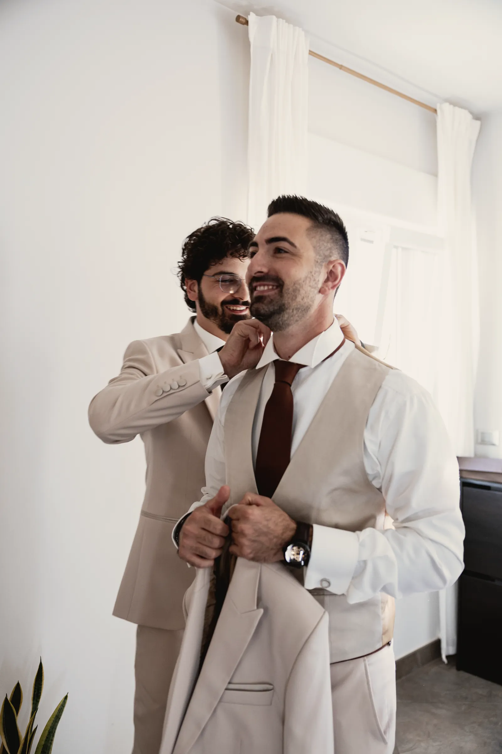 Un uomo in abito chiaro e cravatta ruggine si fa aiutare a indossare la giacca.