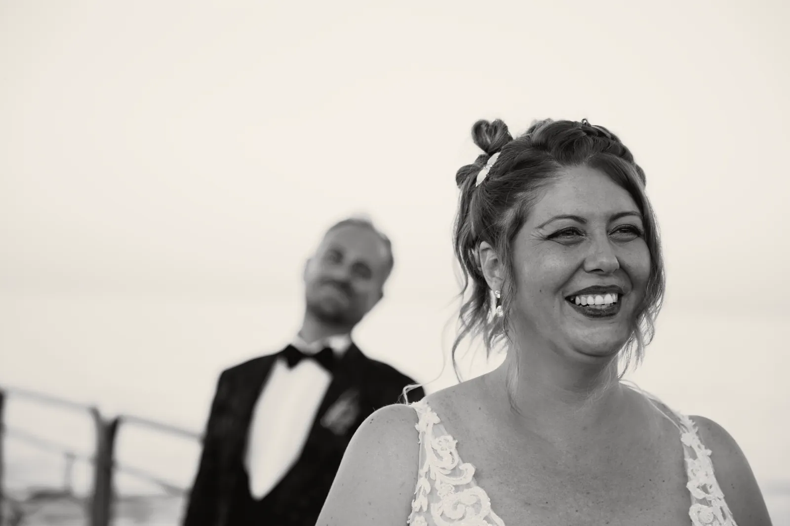 Sul pontile la sposa ride in primo piano; dietro di lei lo sposo, sfocato, piega la testa verso di lei.