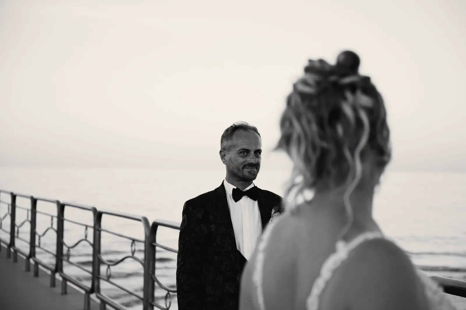 La sposa, di spalle, guarda lo sposo in piedi alla ringhiera del pontile, con il mare dietro.