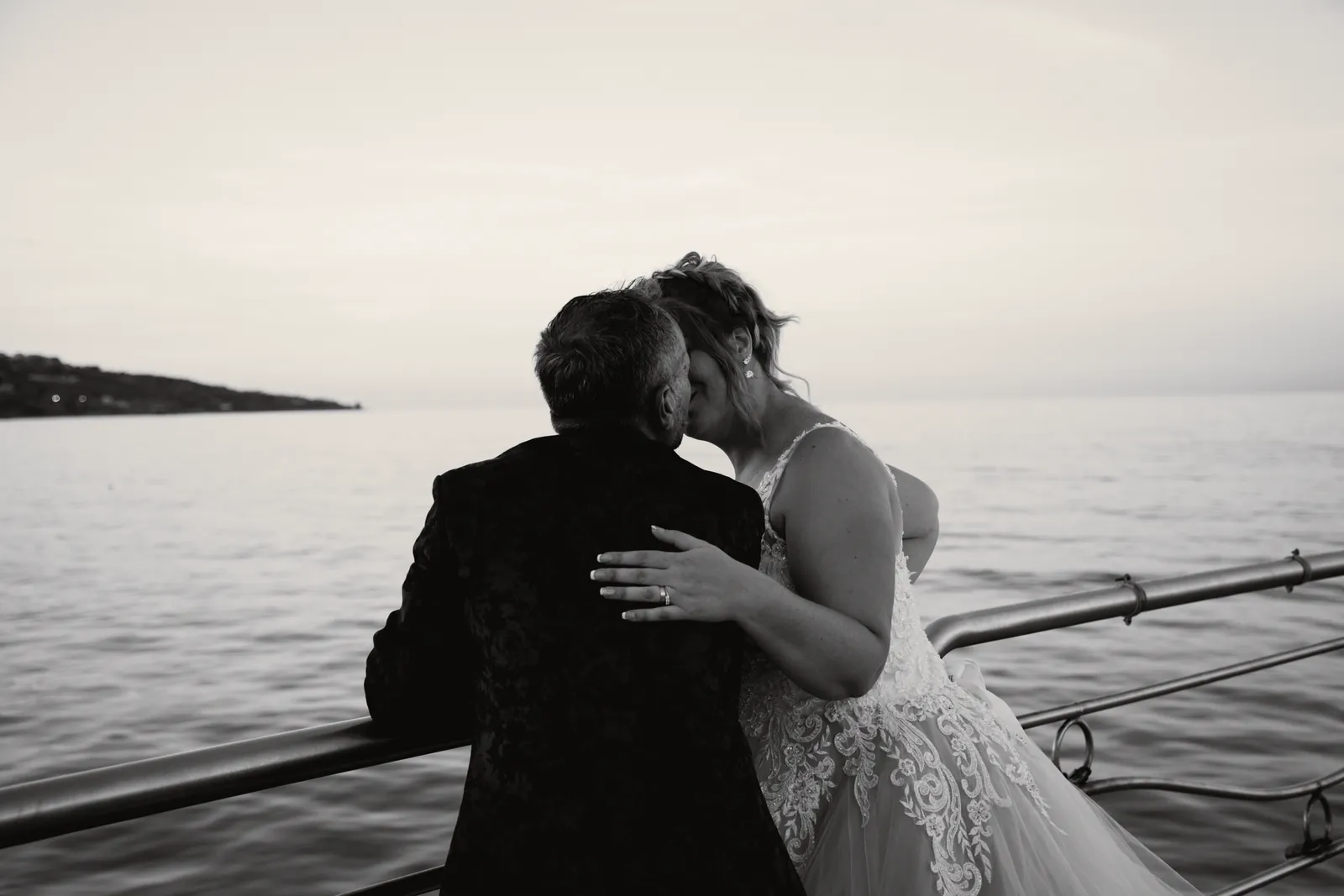 Alla ringhiera del pontile lo sposo, di spalle, bacia la sposa; davanti a loro il mare e il promontorio.
