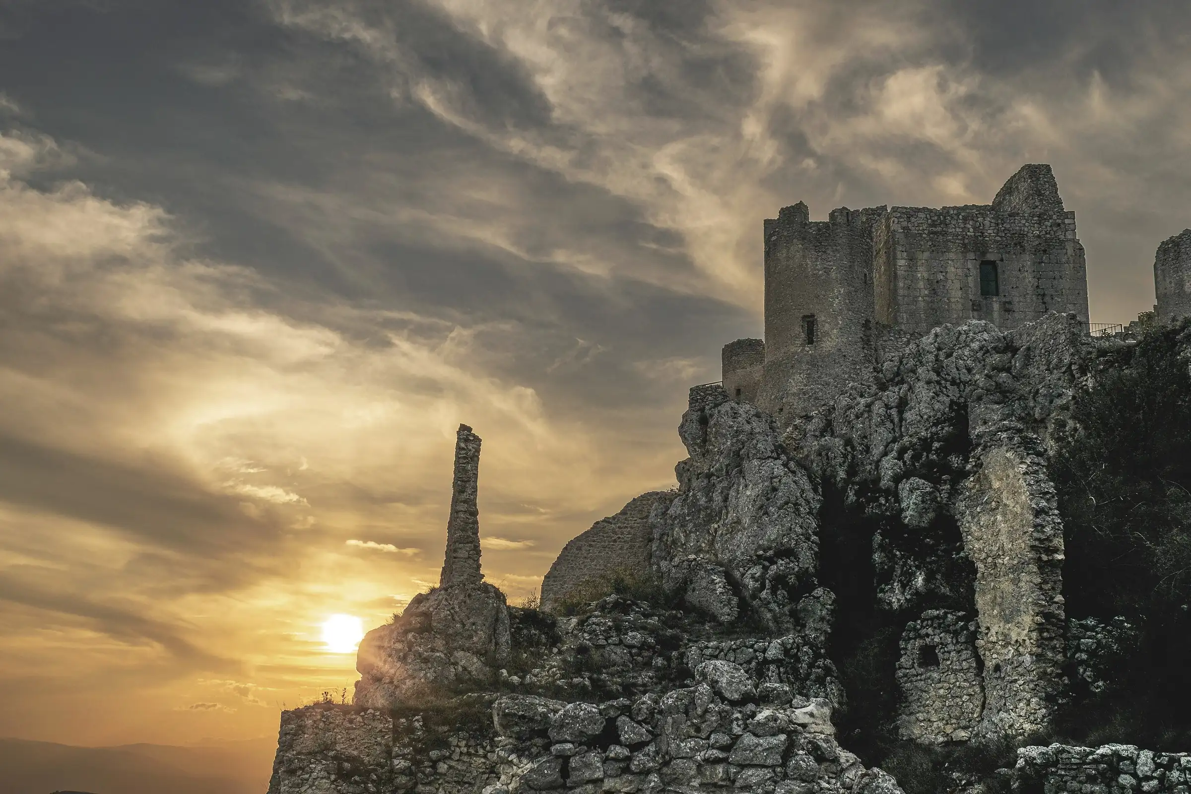 La rocca di Calascio sullo sperone di roccia, controluce al tramonto, con il sole basso sotto le nuvole.