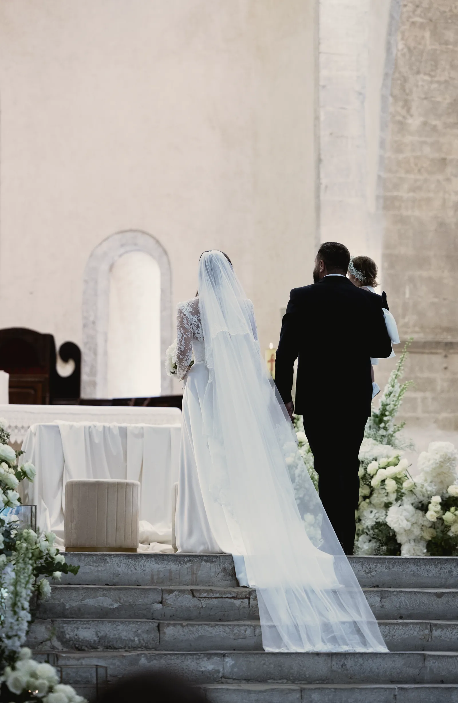 Gli sposi, di spalle, salgono i gradini dell’altare; il velo si allunga sulla pietra.