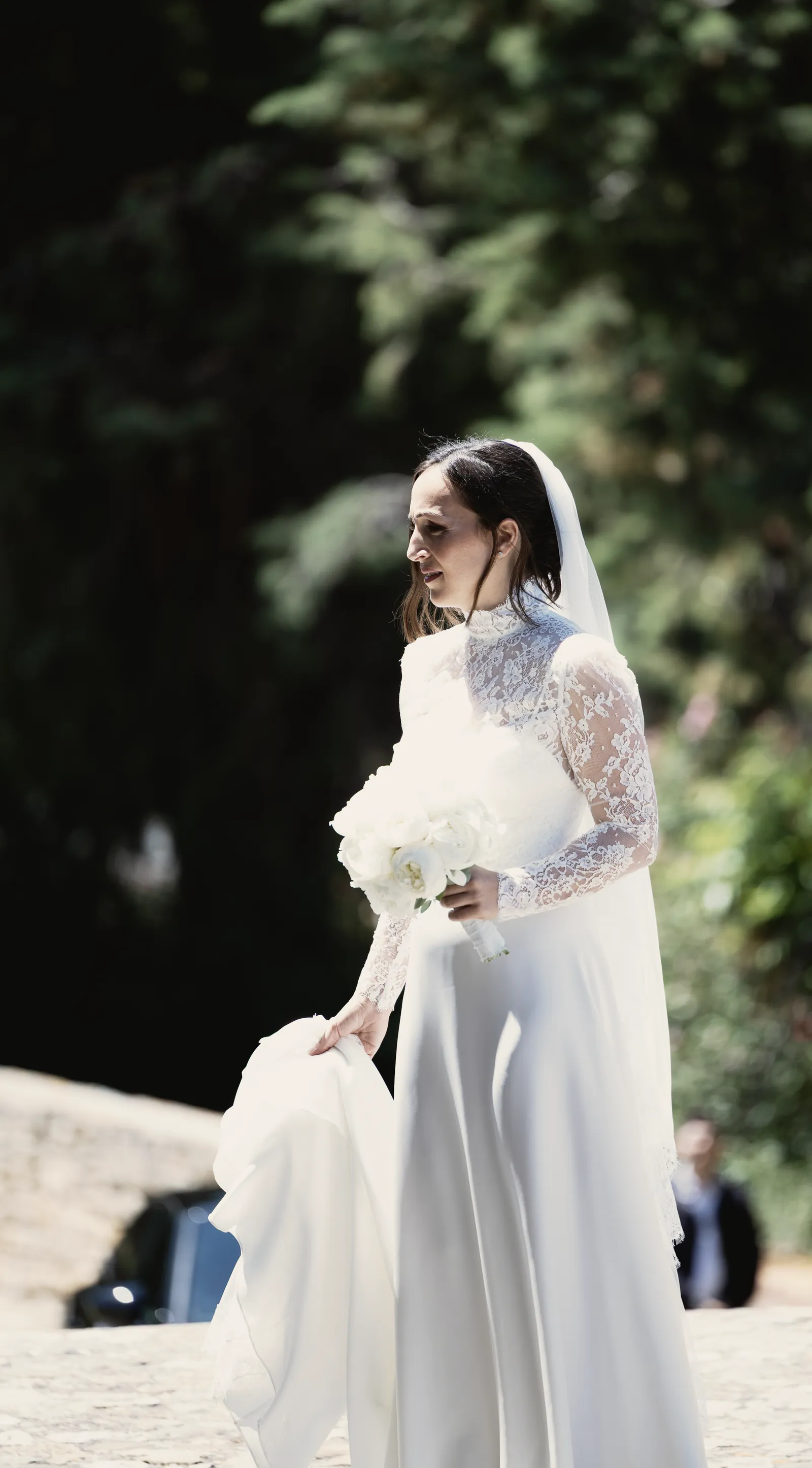 La sposa di profilo fra i pini, il bouquet in una mano e lo strascico nell’altra.